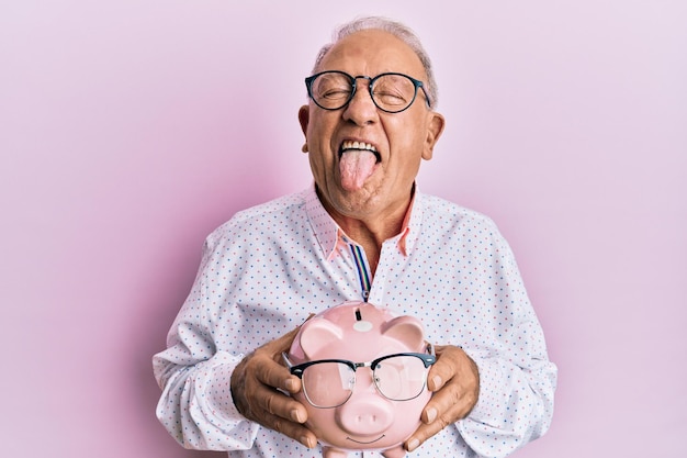 Homem caucasiano sênior segurando o cofrinho com óculos enfiando a língua feliz com expressão engraçada.