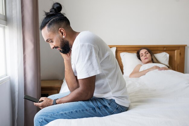 Homem checando seu telefone na cama ao lado de sua esposa