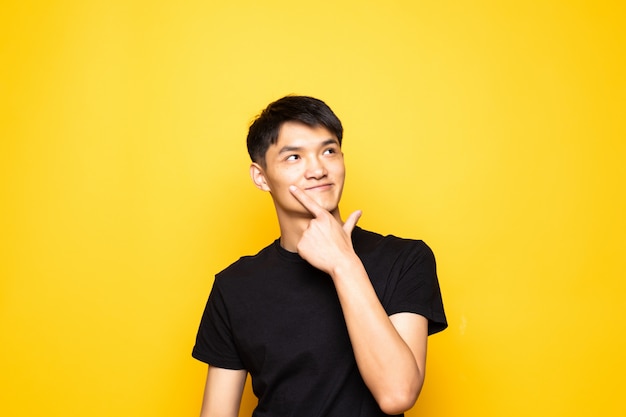 Homem chinês asiático novo com mão no queixo que pensa sobre a pergunta, expressão pensativa que está sobre a parede amarela isolada. Sorrindo com cara de pensativo. Conceito de dúvida.