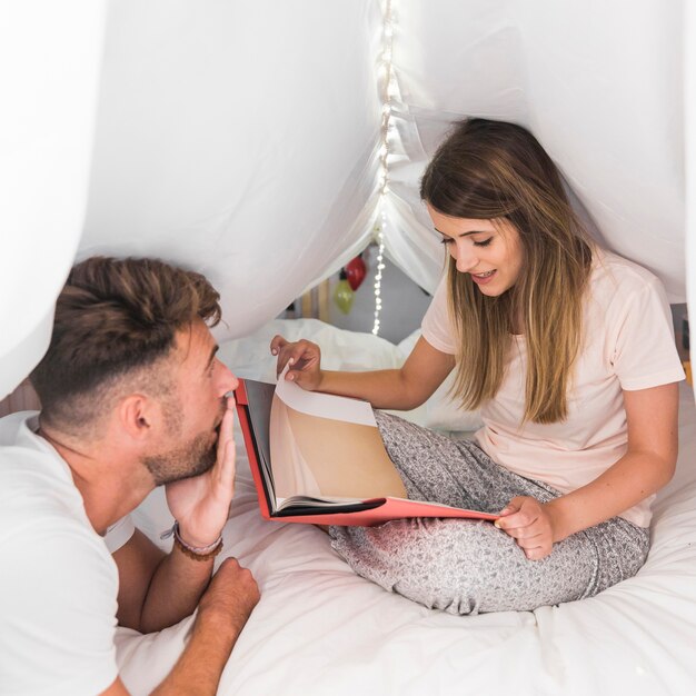Homem chocado olhando mulher segurando o livro sentado na cama sob a cortina