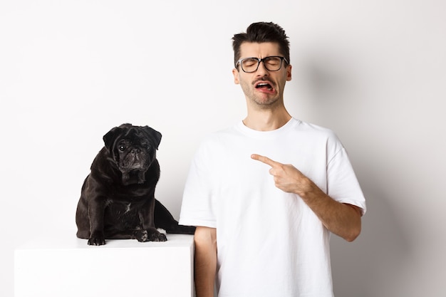 Homem chorando chateado, apontando para um pug preto fofo e soluçando, reclamando de seu animal de estimação, parado triste contra um fundo branco