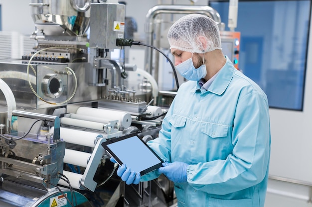 Homem cientista caucasiano em uniforme de laboratório azul perto da máquina de fabricação com eixos mostra tablet vazio perto da imagem foco no tablet olhar para o tablet