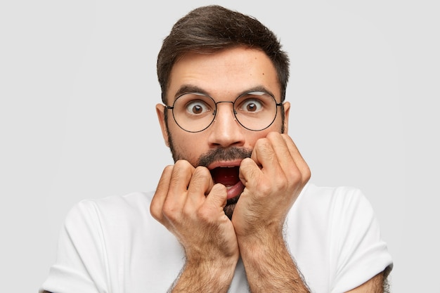 Homem com a barba por fazer assustado morde os punhos nervosamente, abre os olhos amplamente, sente-se ansioso, tem medo de algo apavorado, usa óculos redondos e camiseta branca