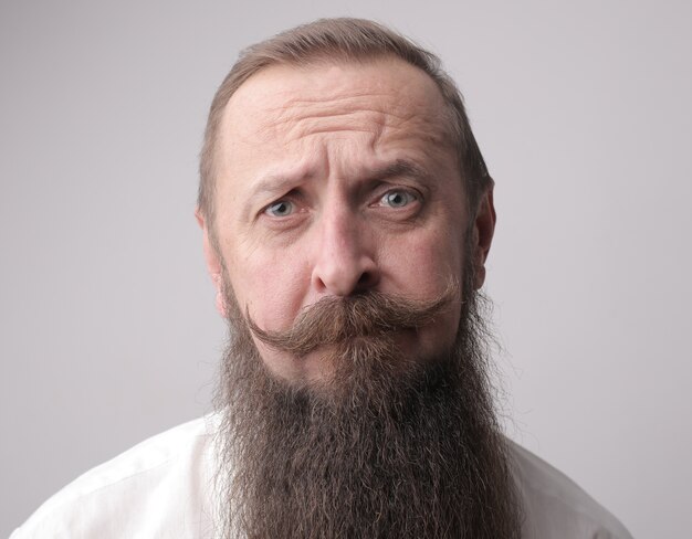 Homem com barba comprida e bigode franzindo a testa em frente a uma parede cinza