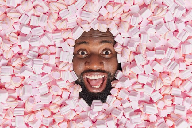 homem com barba espessa parece feliz mantém a boca aberta Afogado em um doce e apetitoso marshmallow gosta de comer delícias saborosas