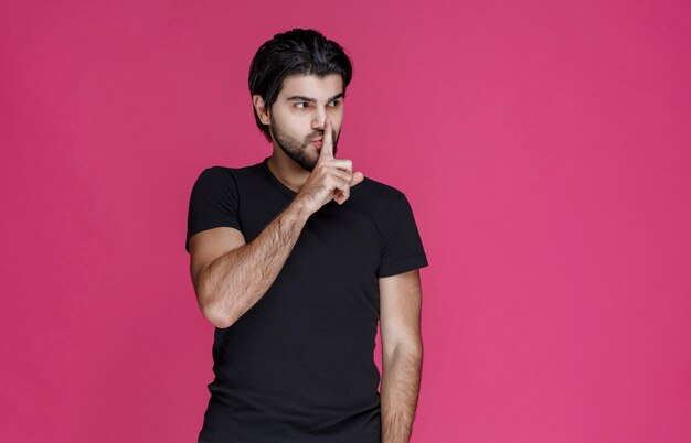 Homem com barba fazendo sinal de silêncio ou apontando alto volume.