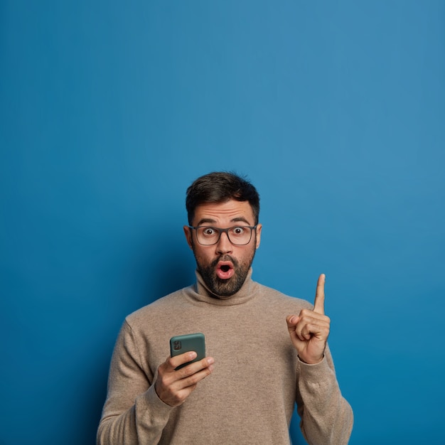 Homem com barba por fazer surpreso segura o telefone, mostra o espaço vazio acima, aponta o dedo indicador, usa óculos e macacão marrom, mantém a boca aberta, isolada sobre fundo azul.