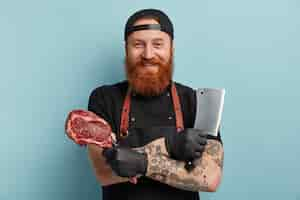 Foto grátis homem com barba ruiva usando avental e luvas segurando uma faca e carne