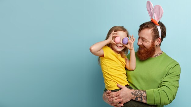 Foto grátis homem com barba ruiva vestindo roupas coloridas e orelhas de coelho segurando a filha