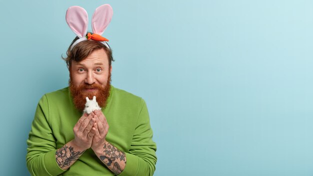 Homem com barba ruiva vestindo roupas coloridas e orelhas de coelho segurando o coelho