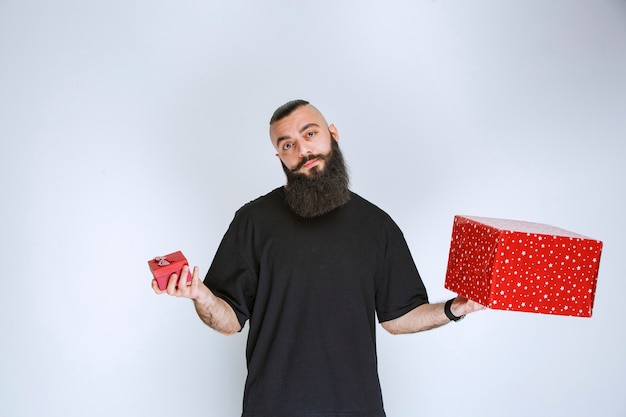 Homem com barba segurando caixas de presente vermelhas e hesitando em fazer a escolha.