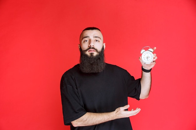 Homem com barba segurando e promovendo um despertador como um produto.