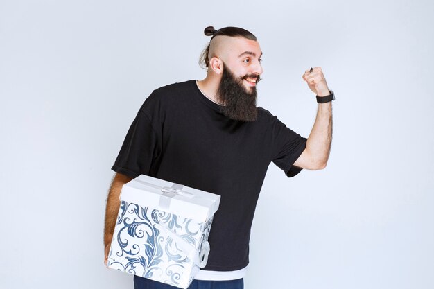 Homem com barba segurando uma caixa de presente azul branca e se sentindo bem-sucedido.