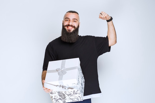 Homem com barba segurando uma caixa de presente azul branca e se sentindo bem-sucedido.