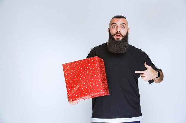 Homem com barba segurando uma caixa de presente vermelha e parece confuso e apavorado.