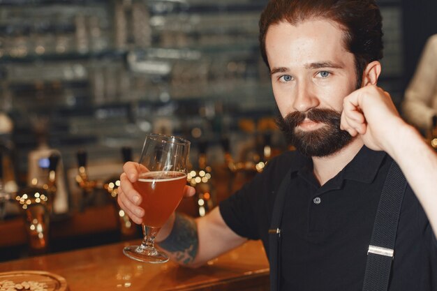 Homem com bigode e barba está no bar e bebe álcool em um copo.