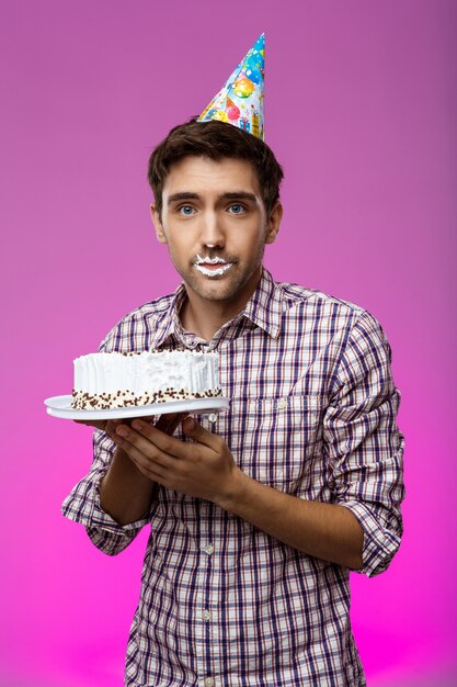 Homem com bolo nos lábios sobre parede roxa. Festa de aniversário.