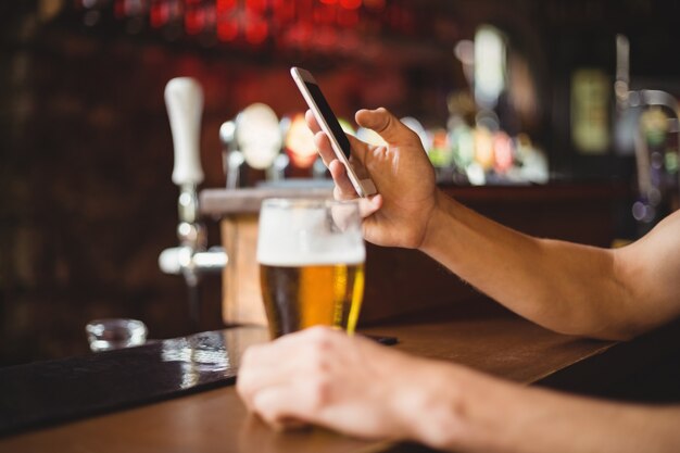 Homem com copo de cerveja, usando telefone celular no balcão