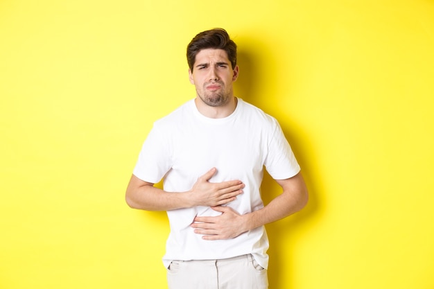 Homem com dor de estômago, fazendo caretas de dor e tocando a barriga, em pé sobre um fundo amarelo.