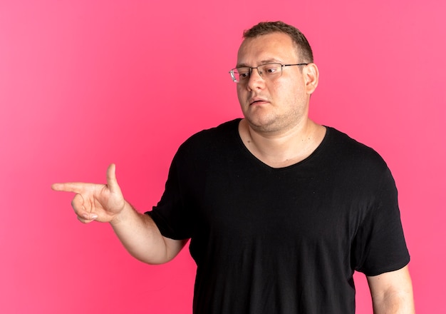 Homem com excesso de peso de óculos, usando camiseta preta, olhando para o lado com o rosto sério apontando com o dedo para o lado sobre rosa