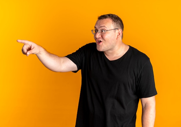 Homem com excesso de peso de óculos, usando uma camiseta preta, apontando com o dedo para algo feliz e animado em pé sobre uma parede laranja