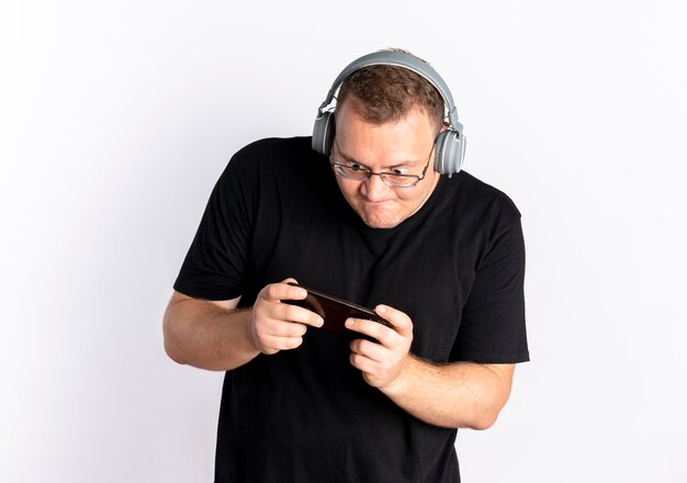 Homem com excesso de peso de óculos, usando uma camiseta preta com fones de ouvido, jogando em seu smartphone em pé sobre uma parede branca