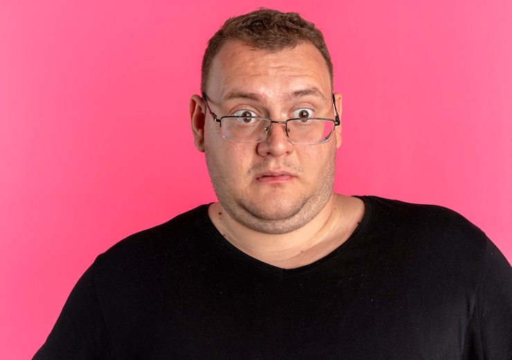 Foto grátis homem com excesso de peso de óculos, usando uma camiseta preta, parecendo de lado surpreso e surpreso em pé sobre uma parede rosa