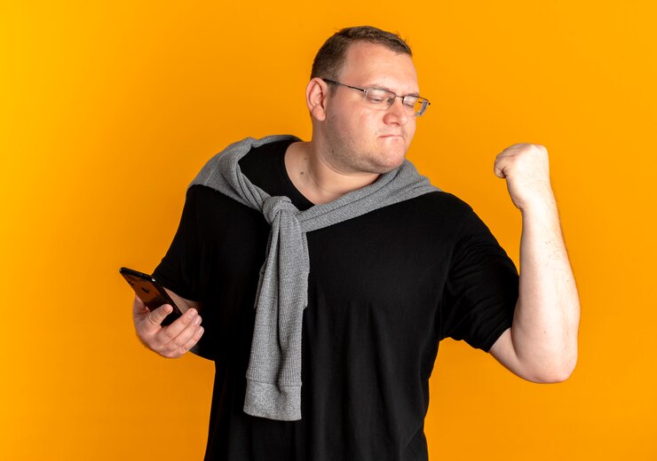 Foto grátis homem com excesso de peso de óculos, vestindo uma camiseta preta, segurando o punho do smartphone, comemorando seu sucesso com o laranja
