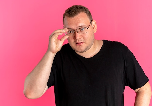 Homem com excesso de peso usando óculos e camiseta preta olhando para algo confuso sobre rosa