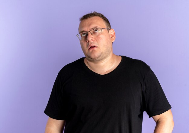 Homem com excesso de peso usando óculos e camiseta preta parecendo cansado e entediado revirando os olhos sobre o azul