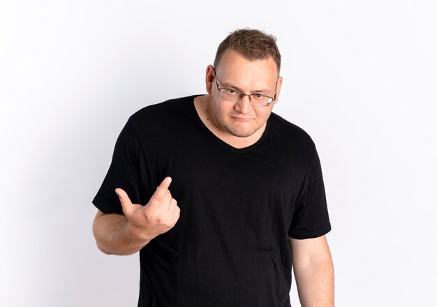 Homem com excesso de peso vestindo uma camiseta preta com óculos, parecendo confuso, mostrando o dedo indicador em pé sobre uma parede branca