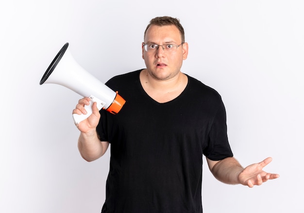 Foto grátis homem com excesso de peso vestindo uma camiseta preta de óculos segurando um megafone, parecendo confuso e descontente em pé sobre uma parede branca