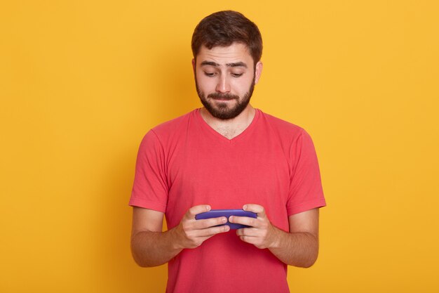 homem com expressão calmafacial, camisa vermelha casual de dreses, jogando jogos online em smartphone ou verificando sua rede social, posando isolado em amarelo. Conceito de tecnologia