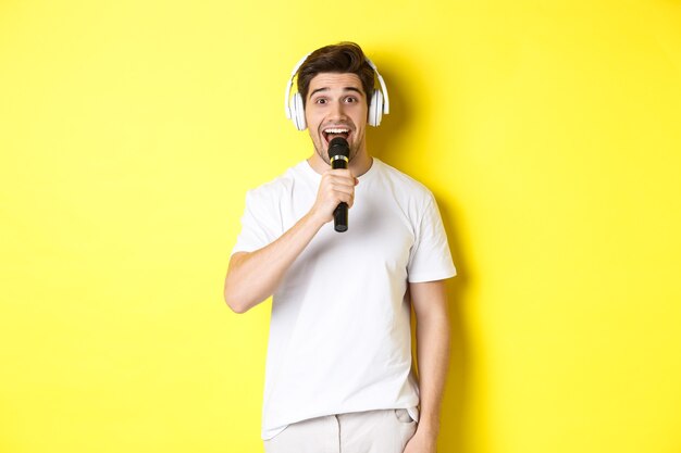 Homem com fones de ouvido segurando um microfone, cantando uma canção de karaokê, em pé sobre um fundo amarelo com roupas brancas