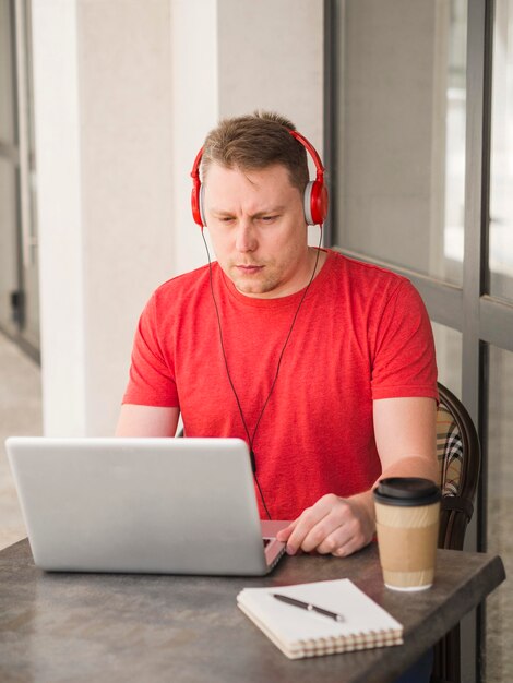 Homem com fones de ouvido, tomando café ao ar livre e trabalhando no laptop