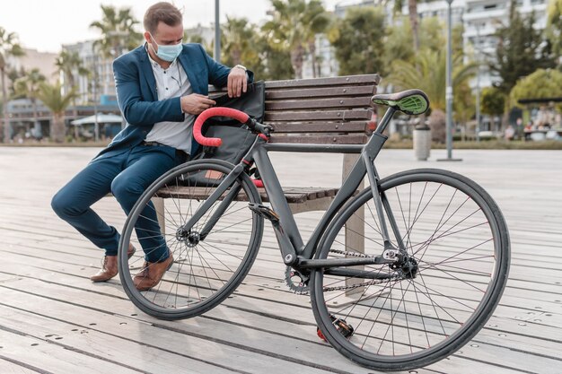 Homem com máscara médica sentado em um banco ao lado de sua bicicleta