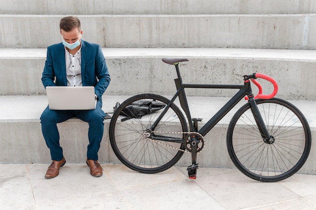 Homem com máscara médica trabalhando em um laptop ao lado de uma bicicleta