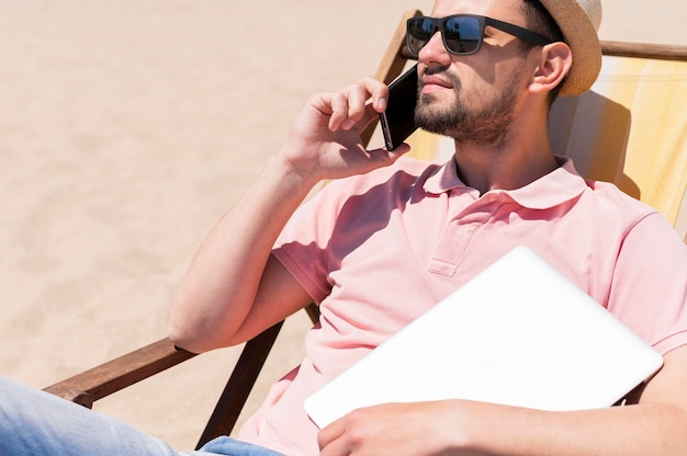 Foto grátis homem com óculos de sol na praia com laptop e smartphone
