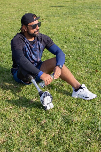 Homem com perna protética relaxante após o treino. Homem caucasiano com barba bebendo água após exercícios, sentado na grama. Esporte, lazer, conceito de deficiência