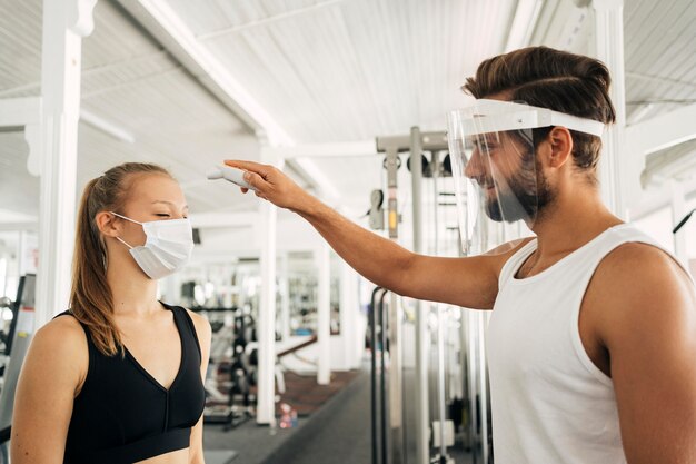 Homem com protetor facial verificando a temperatura da mulher na academia