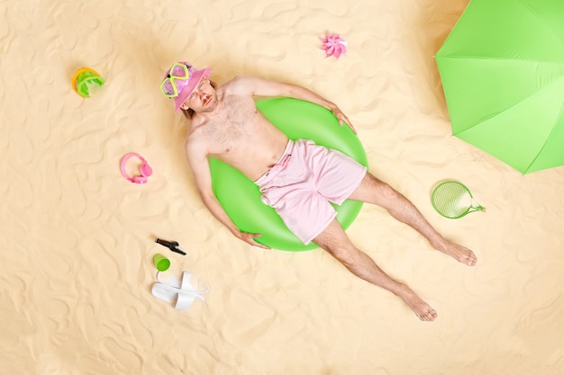 Homem com torso nu deitado em uma pista de natação verde inflada cercado por acessórios de praia poses na areia branca tem expressão de raiva se bronzeando sozinho