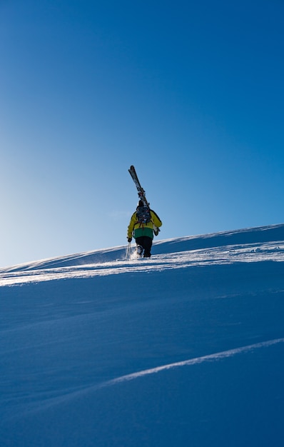 Homem com um casaco amarelo andando na neve carregando uma prancha de esqui