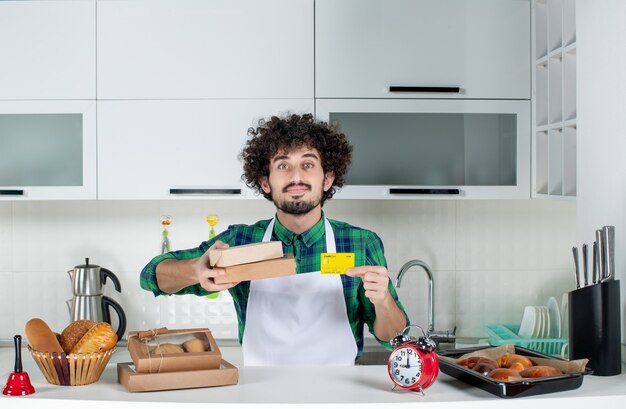 Homem confiante atrás da mesa com vários doces e segurando caixas marrons de cartão de banco na cozinha branca