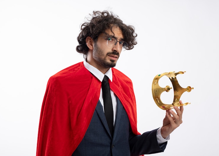 Foto grátis homem confiante e jovem super-herói usando óculos ópticos, usando um terno com capa vermelha, segurando uma coroa