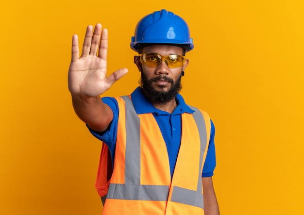 Foto grátis homem confiante jovem construtor afro-americano com óculos de segurança, uniforme com capacete de segurança, gesticulando sinal de pare isolado na parede laranja com espaço de cópia