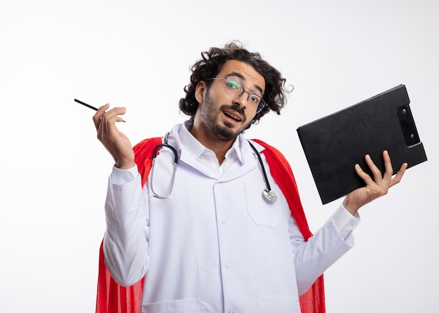 Homem confuso, jovem super-herói caucasiano, usando óculos ópticos, uniforme de médico com capa vermelha e estetoscópio no pescoço, segurando um lápis e uma prancheta isolada na parede branca