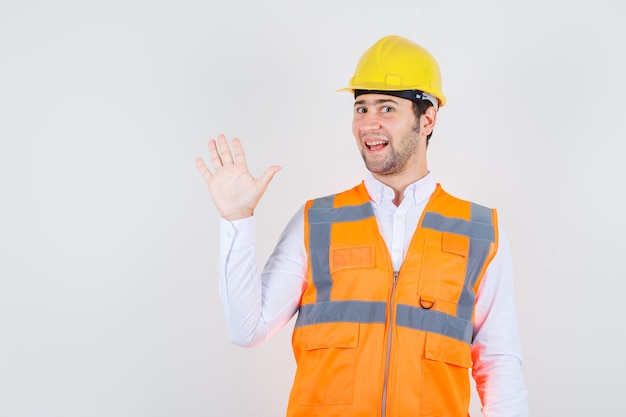 Homem Construtor acenando com a mão para saudação na camisa, uniforme e olhando alegre, vista frontal.