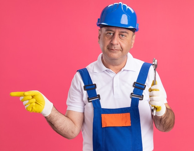 Homem construtor adulto, caucasiano, satisfeito, de uniforme, usando luvas de proteção, segurando a chave inglesa e apontando para o lado na rosa