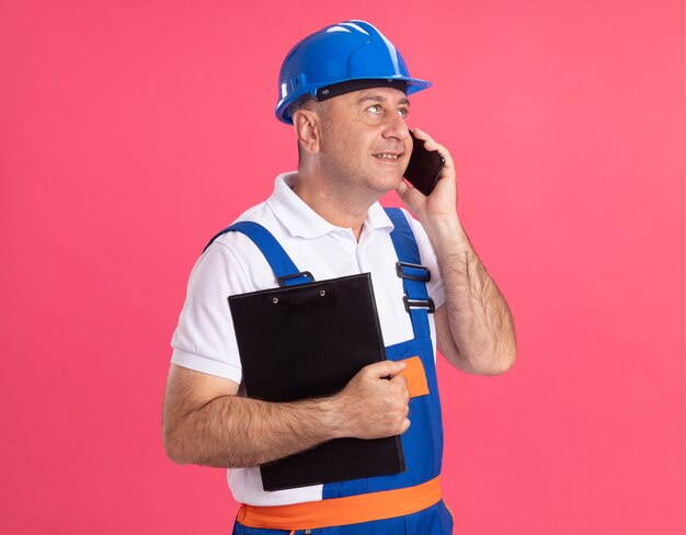 Homem construtor adulto satisfeito, de uniforme, segurando a prancheta e falando ao telefone, olhando para o lado isolado na parede rosa