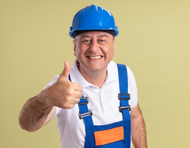 Homem construtor adulto sorridente com uniforme polegar para cima isolado na parede verde oliva